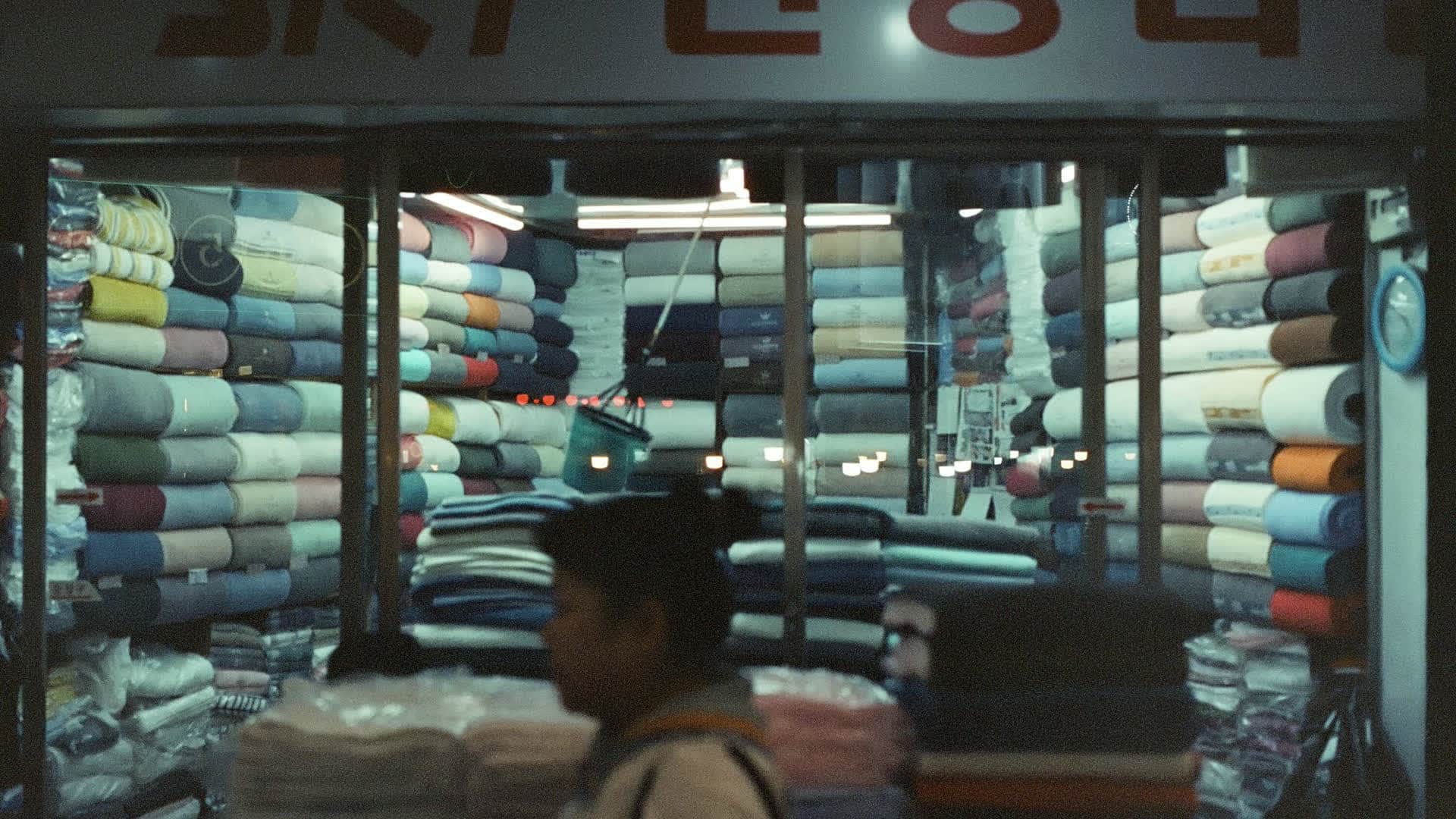 A lit shopfront seen from the street, rolls of cloth stacked inside