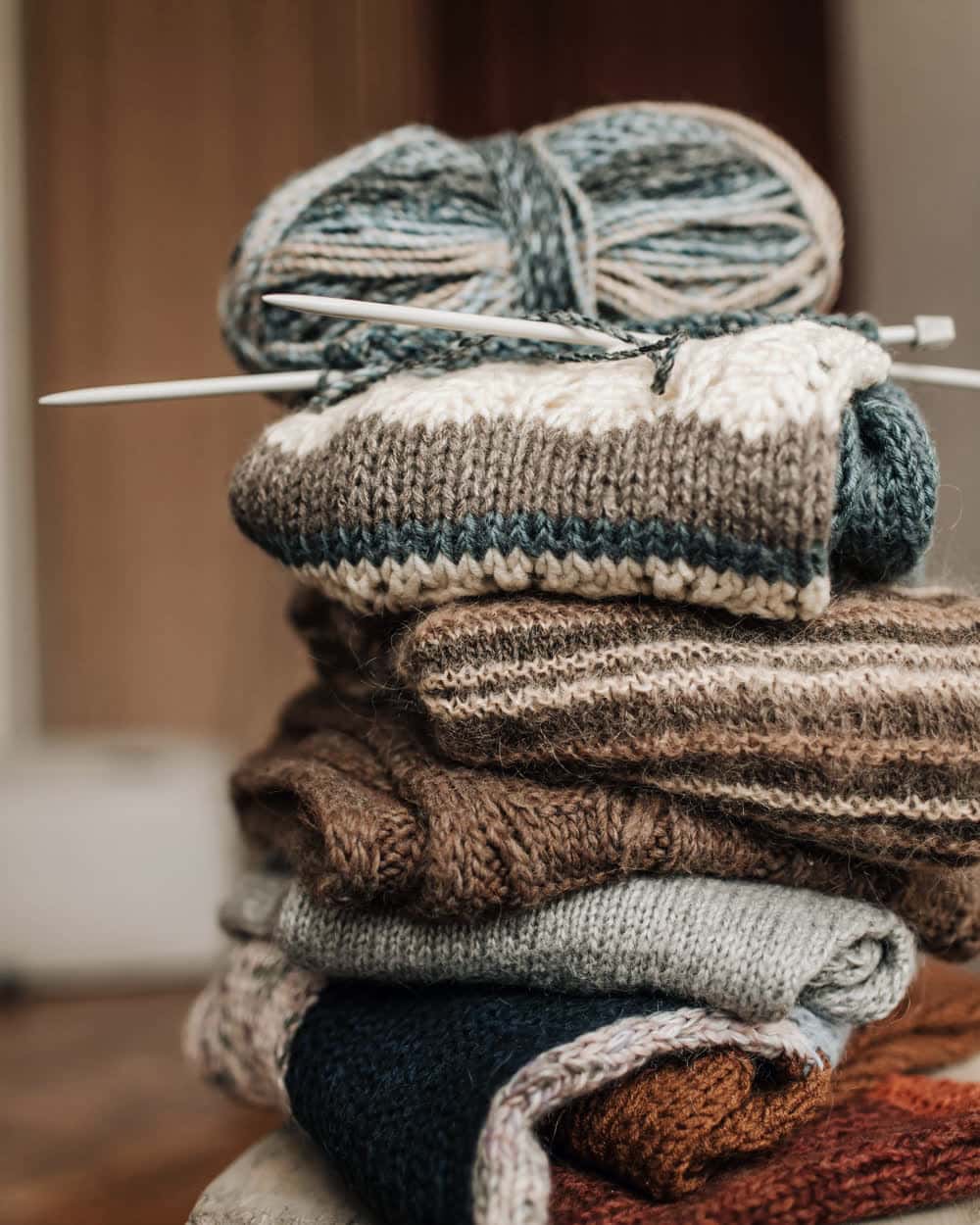 A stack of knitted mufflers in teal, brown, grey and cream, folded on a stool